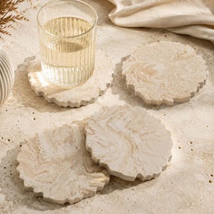 May include: A clear glass of water sits on a decorative coaster. The coaster is a light beige color with a wavy edge and a marbled design. Several coasters are arranged on a textured surface, with a neutral-colored cloth in the background.