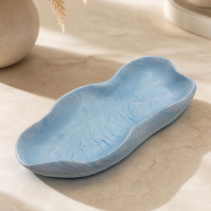 May include: A light blue decorative tray with a marbled effect. The tray has an organic shape and a smooth, curved design. The tray is empty and sits on a light-coloured surface. The tray is likely made of a solid material.
