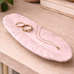 Puede incluir: Una bandeja rosa en forma de pluma contiene dos anillos de oro y un delicado collar de oro con un pequeño colgante. La bandeja está sobre una superficie de madera, mostrando las joyas.