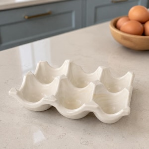 May include: A white ceramic egg tray with six rounded indentations designed to hold individual eggs. The tray features a smooth, glossy finish with subtle cream-coloured marbling throughout its structure, providing a stable base for storage.