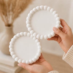 May include: Two white, round coasters with a beaded edge are held in hands. The coasters have a smooth, flat center and a raised, rounded border. The background includes a vase with dried plants.