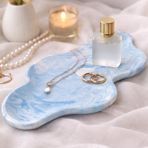 Könnte beinhalten: Ein blau-weiß marmorierter Dekoteller hält eine goldene Halskette mit Anhänger, zwei goldene Ringe und eine kleine Parfümflasche mit goldenem Verschluss. Eine Kerze und eine Vase mit Trockenblumen befinden sich im Hintergrund.