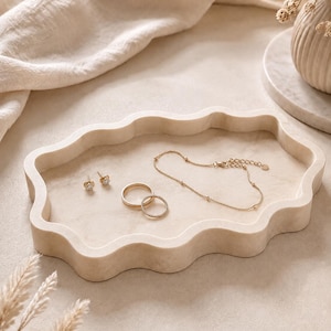 May include: A beige, wavy-edged tray holds jewelry: gold earrings, two rings, and a delicate necklace. The tray sits on a neutral surface, with a soft cloth and a vase of dried flowers in the background. The overall aesthetic is minimalist and elegant.
