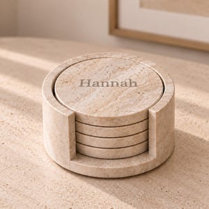 May include: A set of four circular beige marble coasters stacked inside a matching cylindrical marble holder. The top coaster features the name Hannah engraved in a serif font. The stone has a natural veined texture in shades of tan and cream.