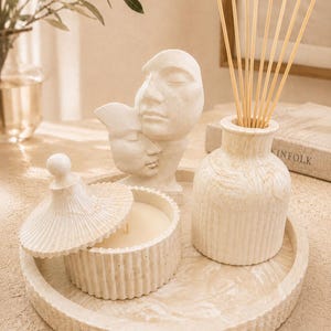 Op de afbeelding: Een crèmekleurig decoratief dienblad met een kaars, een diffuser en een sculptuur van twee gezichten. De kaars zit in een ronde container met een deksel. De diffuser heeft houten stokjes. De sculptuur toont twee gezichten.
