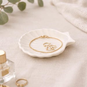 May include: A white, shell-shaped dish displays a gold bracelet and two rings with pearl details. A small perfume bottle and a gold key are in the foreground. The jewellery is presented on a neutral-toned surface.