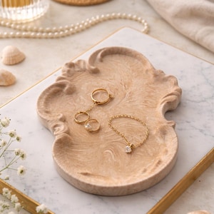 May include: A gold jewellery set, including a ring and bracelet, rests on a decorative, light brown tray. The tray sits on a marble surface with a gold border. Additional accessories include a glass bottle, a candle, and seashells.