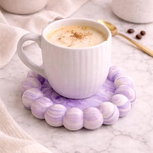 Pode incluir: Uma caneca de cerâmica branca cheia de uma bebida cremosa, polvilhada com canela, repousa sobre um descanso de copo decorativo lavanda e branco. O descanso de copo tem um design único e arredondado. Uma colher dourada e grãos de café estão por perto.