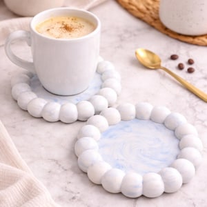 May include: Two white ceramic coasters with a blue marbled design and a beaded edge. One coaster holds a white mug filled with a cappuccino, sprinkled with cinnamon. A gold spoon and coffee beans are nearby.