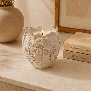 Puede incluir: Una vela encendida en un recipiente crema con un diseño de hojas. La vela está sobre una superficie clara, junto a dos libros titulados "THE REPUBLIC" y "PHAEDO" de Platón. Un jarrón con flores secas está en el fondo.