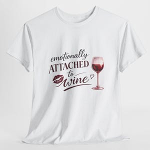 Può includere: T-shirt bianco con la scritta "emotionally ATTACHED to wine" in bordeaux, accompagnata da un'impronta di rossetto e un calice di vino rosso. Il disegno è centrato sulla parte anteriore della maglietta.