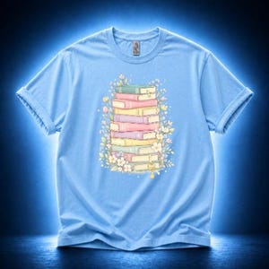 May include: Light blue t-shirt featuring a stack of colorful books with floral accents. The books are in pastel shades of pink, yellow, green, and purple. The design is centered on the shirt, with a crew neck and short sleeves.