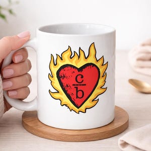 Op de afbeelding: Witte keramische mok met een rood hartontwerp en de letters "c/b" erin, omringd door gele en oranje vlammen. De mok staat op een houten onderzetter. Een hand houdt de mok vast.
