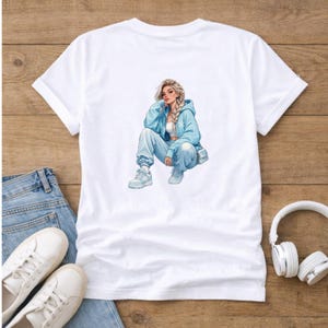 Può includere: T-shirt bianco con un'illustrazione di una donna in tuta blu. La donna bionda indossa una felpa con cappuccio blu, pantaloni della tuta e scarpe da ginnastica. La maglietta è appoggiata su una superficie di legno con jeans, scarpe bianche e cuffie.