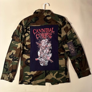 以下が含まれることがあります： 背中に赤い「CANNIBAL CORPSE」のバンド名とグラフィックイラストが描かれた大きなパッチが付いた迷彩ジャケット。右袖には小さなパッチも付いています。白いハンガーに掛けられています。