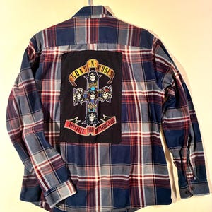 May include: A blue, red, and white plaid flannel shirt with a Guns N' Roses patch on the back. The patch features a cross design with skulls and the words "GUNS N' ROSES" and "APPETITE FOR DESTRUCTION". The shirt is hanging on a white hanger.