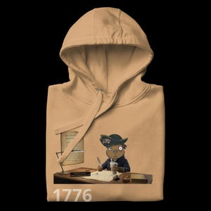 Op de afbeelding: Beige hoodie met een cartoon illustratie van een bever met een steekhoed die op een document schrijft. De tekst "DECLARATION INDEPENDENCE" en "1776" is wit.