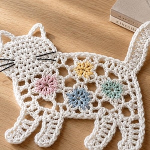 Könnte beinhalten: Ein weißes, gehäkeltes Deckchen in Katzenform mit einem durchbrochenen Muster und vier kleinen, pastellfarbenen Blumenmotiven in Gelb, Rosa, Blau und Mintgrün. Die Katzenfigur hat drei schwarze Garnschnurrhaare auf jeder Gesichtshälfte.