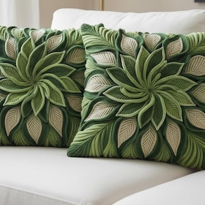 Könnte beinhalten: Zwei quadratische Zierkissen mit einem botanischen Design. Die Kissen weisen ein detailliertes Muster aus grünen Blättern und cremefarbenen Akzenten auf, wodurch ein strukturierter, dreidimensionaler Effekt entsteht. Sie liegen auf einem weißen Sofa, was auf Heimdekoration hindeutet.