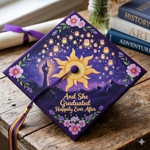 May include: Purple graduation cap with a gold sun design, floating lanterns, and the text "And She Graduated Happily Ever After." The cap is decorated with pink flowers and green leaves. A purple tassel hangs from the cap.