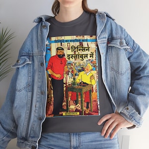 May include: A dark gray t-shirt featuring a graphic print of a comic book cover with two characters, one in a red sweater and one in a yellow sweater, sitting at a table. The text on the graphic includes Hindi script and the word Casterman.