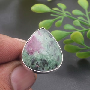 Könnte beinhalten: Ein tropfenförmiger Ring mit einem polierten Zoisit-Stein, der deutliche Einschlüsse von magentafarbenem Rubin und dunklen schwarzen Mineralflecken aufweist und in einer silberfarbenen Metallfassung sitzt.