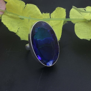 Könnte beinhalten: Ein silberfarbener Ring mit einem großen, ovalen, facettierten tiefblauen Edelstein, der in einer glatten Metallfassung sitzt. Der Stein zeigt eine reflektierende Oberfläche mit verschiedenen Blau- und Dunkeltönen und ist in einer einfachen Ringschiene gefasst.