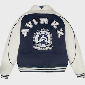 May include: A varsity-style jacket with a cream-colored body and navy blue accents. The back features a large emblem with the text "AVIREX" and "SINCE 1975 NEW YORK CLASSIC". The jacket has striped cuffs and a collar.