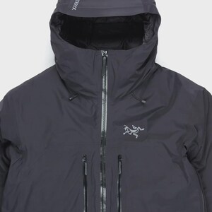 Puede incluir: Chaqueta con capucha gris oscuro con cremallera completa y m&uacute;ltiples bolsillos con cremallera. La chaqueta presenta el nombre de la marca "Arc'teryx" en la capucha y un logotipo en el pecho. La chaqueta parece estar hecha de un material impermeable.