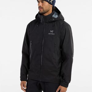 May include: A black hooded jacket with the Arc'teryx logo and a navy blue beanie with the word "TERYX" in white. The jacket has a full-length zipper and multiple pockets. The model is wearing black pants.