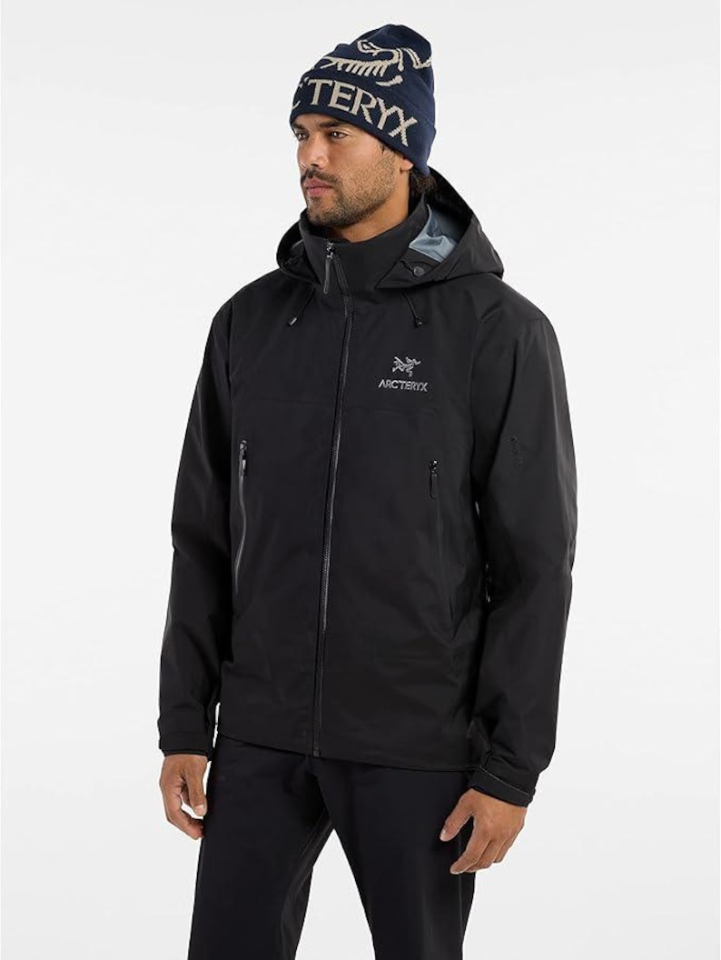 May include: A black hooded jacket with the Arc'teryx logo and a navy blue beanie with the word "TERYX" in white. The jacket has a full-length zipper and multiple pockets. The model is wearing black pants.