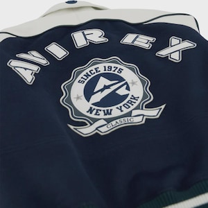 May include: Navy blue and white varsity jacket with the text "AIREX" across the back. The jacket features a patch with the text "SINCE 1975 NEW YORK CLASSIC". The collar is white, and the jacket has a button closure.
