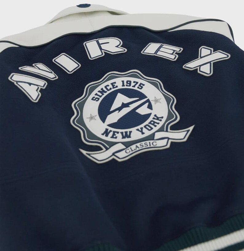 May include: Navy blue and white varsity jacket with the text "AIREX" across the back. The jacket features a patch with the text "SINCE 1975 NEW YORK CLASSIC". The collar is white, and the jacket has a button closure.