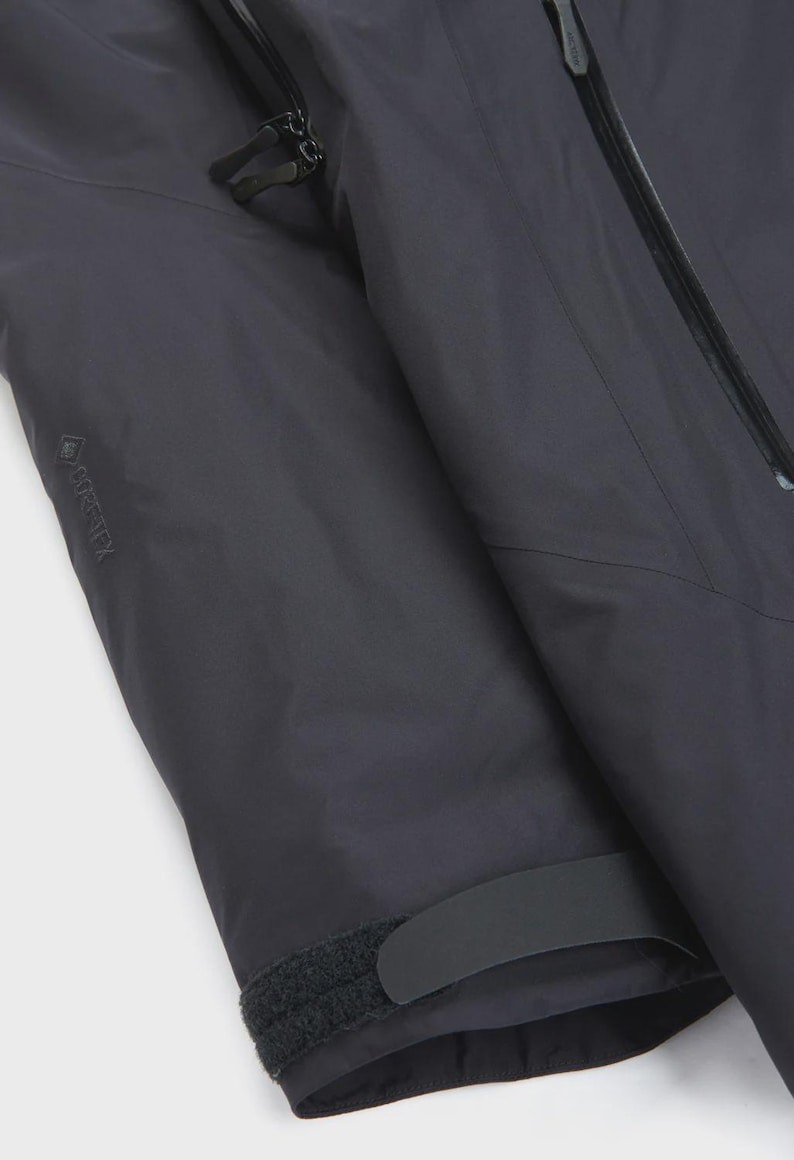 Puede incluir: Primer plano de una manga de chaqueta gris oscuro. La manga presenta una cremallera negra, un pu&ntilde;o de velcro y el logotipo GORE-TEX. La chaqueta parece estar hecha de un material duradero e impermeable, adecuado para actividades al aire libre.