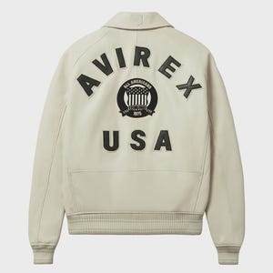 Op de afbeelding: Cr&egrave;mekleurige leren jas met het woord "AVIREX" in zwarte letters op de rug. Daaronder een cirkelvormig embleem met de woorden "ALL AMERICANS" en "USA" in zwart.