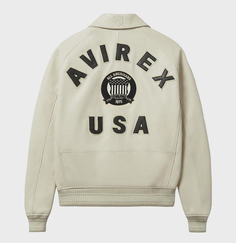 Op de afbeelding: Cr&egrave;mekleurige leren jas met het woord "AVIREX" in zwarte letters op de rug. Daaronder een cirkelvormig embleem met de woorden "ALL AMERICANS" en "USA" in zwart.