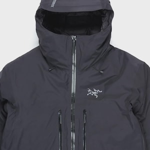 Puede incluir: Chaqueta con capucha gris oscuro con cremallera completa y m&uacute;ltiples bolsillos con cremallera. La chaqueta presenta la marca "ARCTERYX" en la capucha y un logotipo en el pecho. La chaqueta parece estar hecha de un material impermeable.