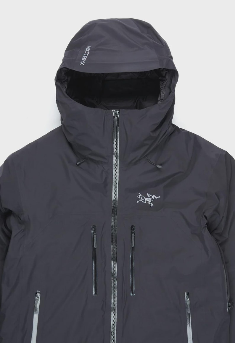 Puede incluir: Chaqueta con capucha gris oscuro con cremallera completa y m&uacute;ltiples bolsillos con cremallera. La chaqueta presenta la marca "ARCTERYX" en la capucha y un logotipo en el pecho. La chaqueta parece estar hecha de un material impermeable.