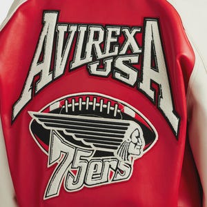 May include: Close-up of the back of a red and white varsity jacket. The jacket features a large embroidered patch with the words "AVIREX US A" in white, along with a football and a Native American head graphic. The number "75ers" is also displayed.