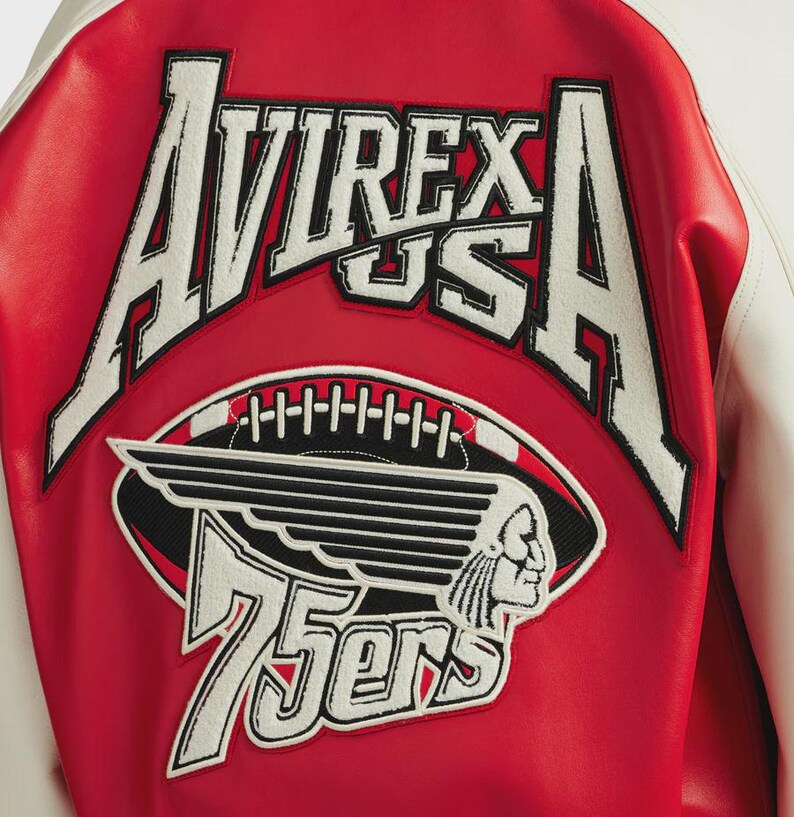 May include: Close-up of the back of a red and white varsity jacket. The jacket features a large embroidered patch with the words "AVIREX US A" in white, along with a football and a Native American head graphic. The number "75ers" is also displayed.