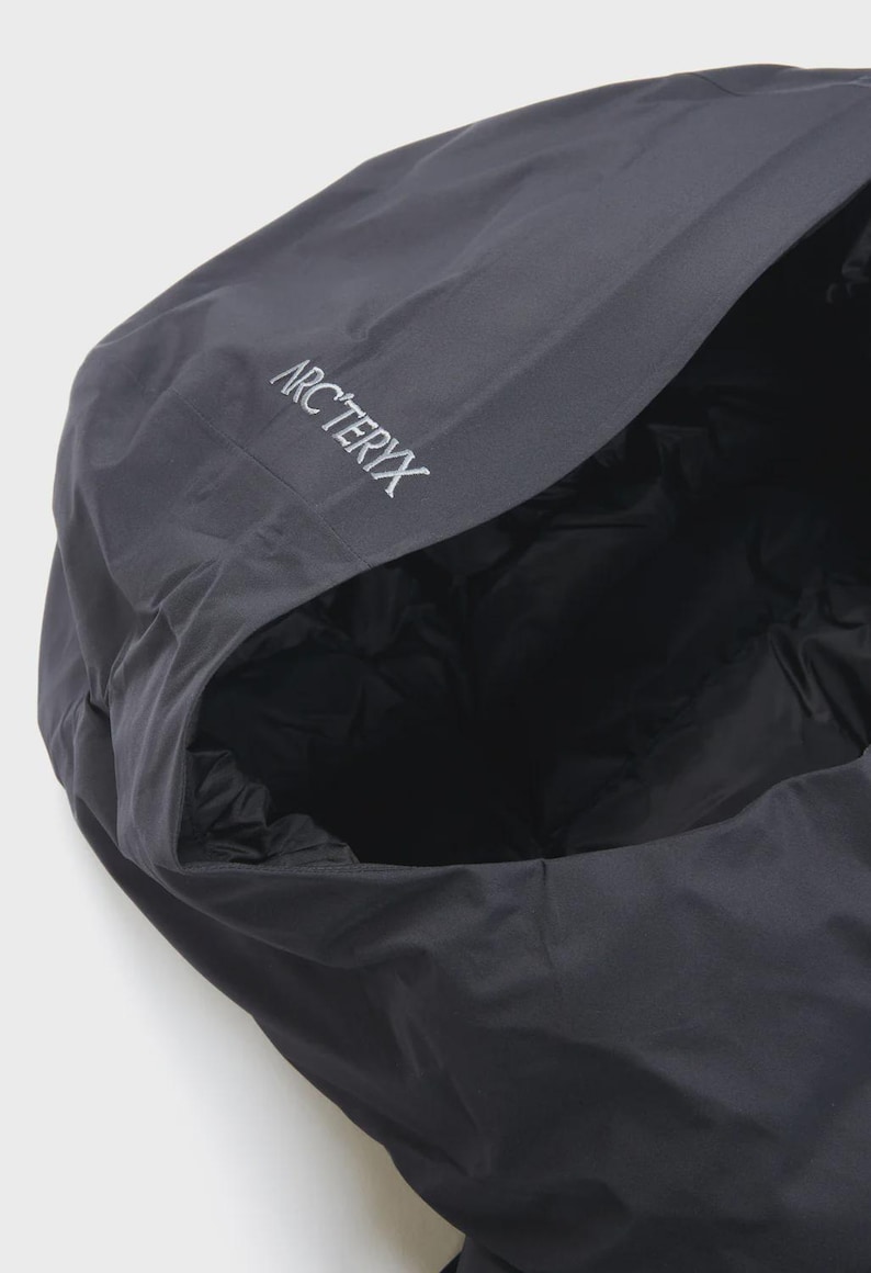 Puede incluir: Primer plano de una chaqueta con capucha gris oscuro con el nombre de la marca "Arc'teryx" bordado en blanco en la capucha. El interior de la capucha es negro y acolchado. La chaqueta parece estar hecha de un material duradero y resistente a la intemperie.