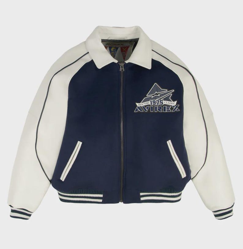 May include: A navy blue and white varsity jacket with a zip-up front. The jacket features white sleeves, a white collar, and a striped hem and cuffs. The chest displays the text "AVIREX 1975 CLASSIC" with a mountain graphic.