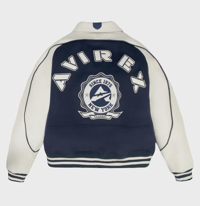 May include: A varsity-style jacket with a cream-colored body and navy blue accents. The back features a large emblem with the text "AVIREX" and "SINCE 1975 NEW YORK CLASSIC". The jacket has striped cuffs and a collar.