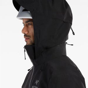 May include: A black hooded jacket with a high collar and a zipper closure. The jacket has a drawstring hood and the Arc'teryx logo on the chest. The person is wearing a white helmet under the hood.