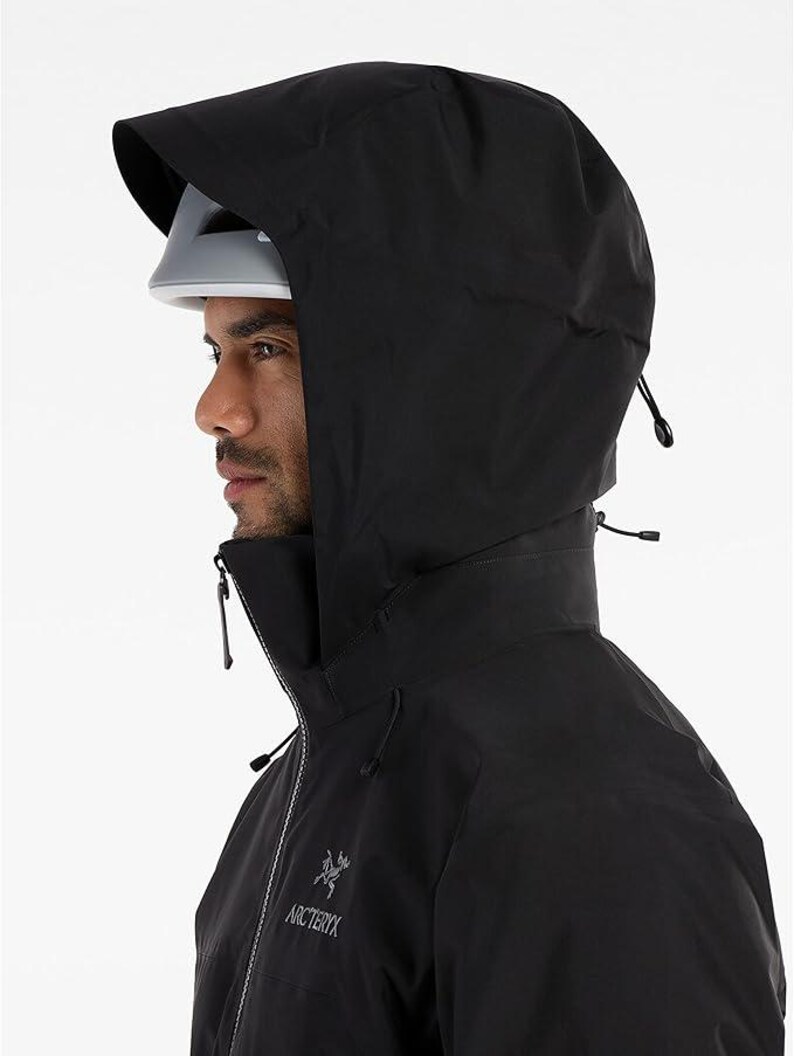 May include: A black hooded jacket with a high collar and a zipper closure. The jacket has a drawstring hood and the Arc'teryx logo on the chest. The person is wearing a white helmet under the hood.