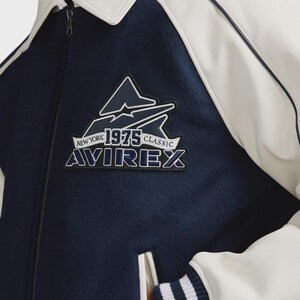 May include: A navy blue and white jacket with the text "NEW YORK 1975 CLASSIC AVIREX" on a patch. The jacket has a white collar and white accents. The jacket is a classic style.
