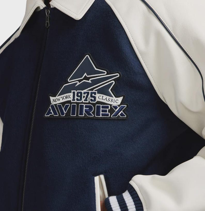 May include: A navy blue and white jacket with the text "NEW YORK 1975 CLASSIC AVIREX" on a patch. The jacket has a white collar and white accents. The jacket is a classic style.