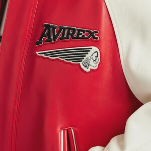 May include: Red leather jacket with white sleeves. The jacket features the embroidered text "AVIREX" above a patch with a Native American head and wing design. The jacket has a classic varsity style.