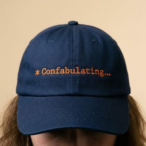 May include: A navy blue baseball cap with the embroidered text "* Confabulating..." in orange. The cap has a curved brim and a button on top. The hat is made of a soft fabric.