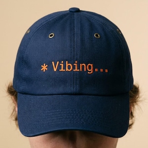 May include: Navy blue baseball cap with the word "Vibing..." embroidered in orange. The cap has a curved brim, a button on top, and adjustable strap. A casual accessory for everyday wear.